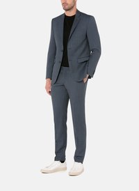 Mand iført en skræddersyet blå jakkesæt med en sort skjorte og hvide sneakers, stående med den ene hånd i lommen mod en ensfarvet baggrund.