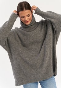 Szary, oversize sweter z golfem o prążkowanej strukturze; posiada szerokie rękawy i luźny fason, stylizowany z niebieskimi dżinsami.