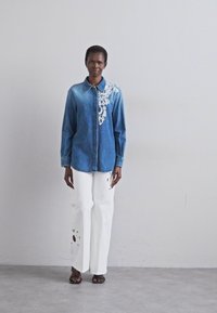 Camicia in denim con un motivo ricamato bianco sulla spalla sinistra, abbinata a pantaloni bianchi a gamba larga con dettagli strappati.