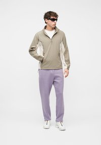 Jeune homme portant une veste beige à fermeture à glissière partielle, un pantalon lavande, des baskets blanches et des lunettes de soleil noires, debout avec une main dans la poche.