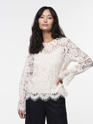 Blouse en dentelle couleur blanc cassé avec des manches longues, un ourlet festonné avec des franges et des motifs floraux. Portée avec un pantalon sombre.