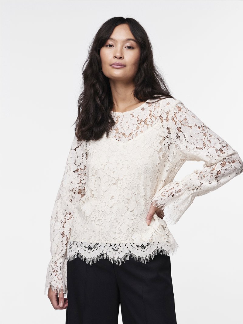 Kanten blouse in off-white met lange mouwen, een gegolfde zoom met franjes en bloempatronen. G gedragen met donkere broeken.