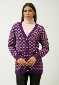 Lila Strickjacke mit kontrastierendem hellrosa Logo-Muster, V-Ausschnitt, langen Ärmeln, Frontknöpfen und Seitentaschen, kombiniert mit einem rosa Rollkragenpullover.