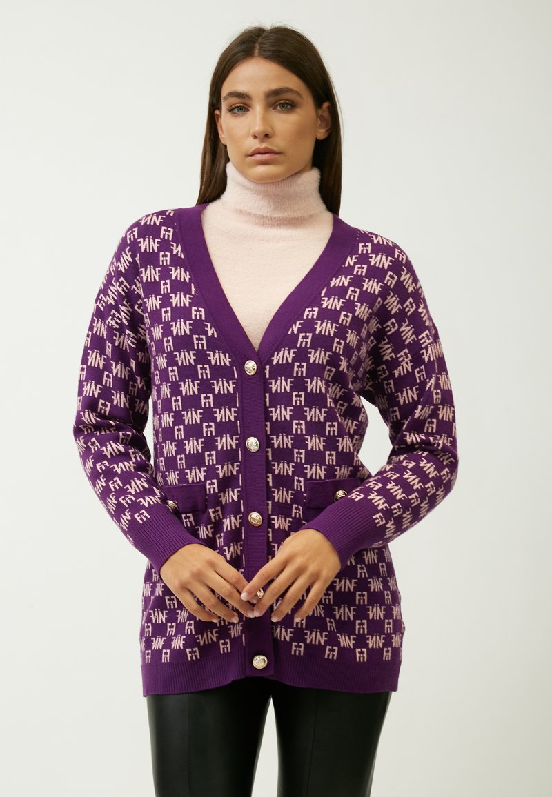 Lila Strickjacke mit kontrastierendem hellrosa Logo-Muster, V-Ausschnitt, langen Ärmeln, Frontknöpfen und Seitentaschen, kombiniert mit einem rosa Rollkragenpullover.