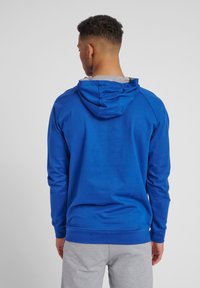 Blå huvtröja med grått foder, med lång ärm och ribbat avslut, mot en enfärgad bakgrund.