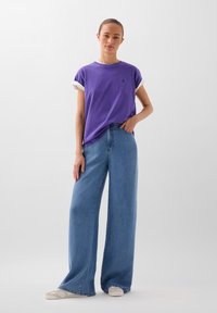 T-shirt en coton violet avec des poignets roulés blancs, associé à un jean large bleu clair. Le tissu semble doux, sans motifs ni accents visibles.