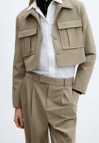 Veste beige cropped avec deux grandes poches avant, col plat et fermeture à boutons, associée à un pantalon beige taille haute et une chemise blanche.
