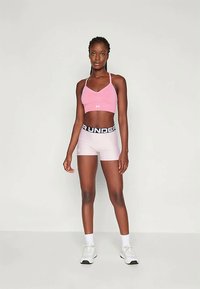 Rosa sport bh og lyse rosa shorts med en svart "Under Armour" elastisk kant, designet for treningstøy. Modellen står foran en nøytral bakgrunn.