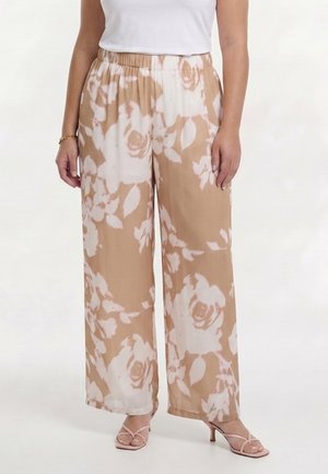 Kvinde iført beige bukser med vide ben med store hvide blomsterprint, kombineret med en hvid ærmeløs top og beige hælede sandaler.