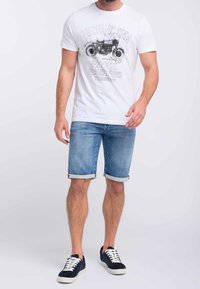 Bílý grafický tričko s černým potiskem motorky, světle modré džíny šortky s ohrnutými lemy, a tmavě modré tenisky s bílou podrážkou.