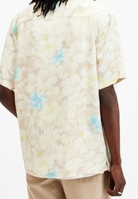 Uomo che indossa una camicia beige a maniche corte con un motivo floreale bianco e azzurro chiaro, abbinata a pantaloni beige, visto da dietro.