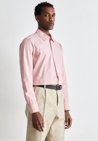Camicia button-up rosa chiaro con colletto, maniche lunghe e polsini a strappo, abbinata a pantaloni beige texturizzati. Cintura nera in evidenza.