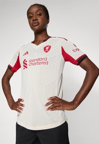 Bela kratka majica s kratkimi rokavi z rdečimi detajli, z v-izrezom, heksagonalnim vzorcem in logotipi podjetij Standard Chartered in Adidas.
