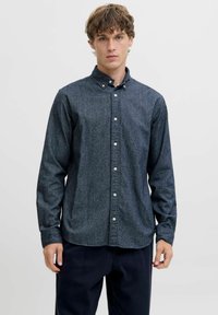 Jauns vīrietis ar cirtainiem matiem, tērpies tumši pelēkā krekla ar pogām un tumšās biksēs, stāv uz vienkārša gaiša fona.