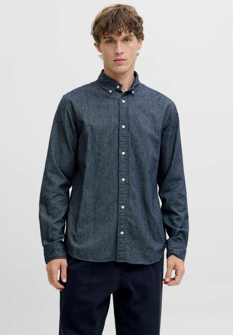 Jauns vīrietis ar cirtainiem matiem, tērpies tumši pelēkā krekla ar pogām un tumšās biksēs, stāv uz vienkārša gaiša fona.