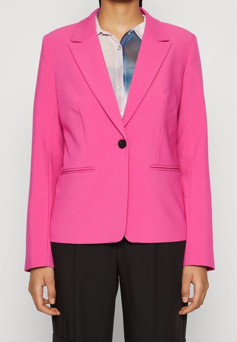 Personne portant un blazer rose vif à un seul bouton par-dessus une chemise blanche et bleue à boutons, avec un pantalon noir visible de la taille vers le bas.