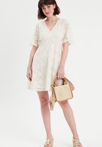 Robe en dentelle blanc cassé avec un col en V, des manches courtes et une silhouette en A. Le mannequin tient un petit sac tissé avec une sangle à motifs.