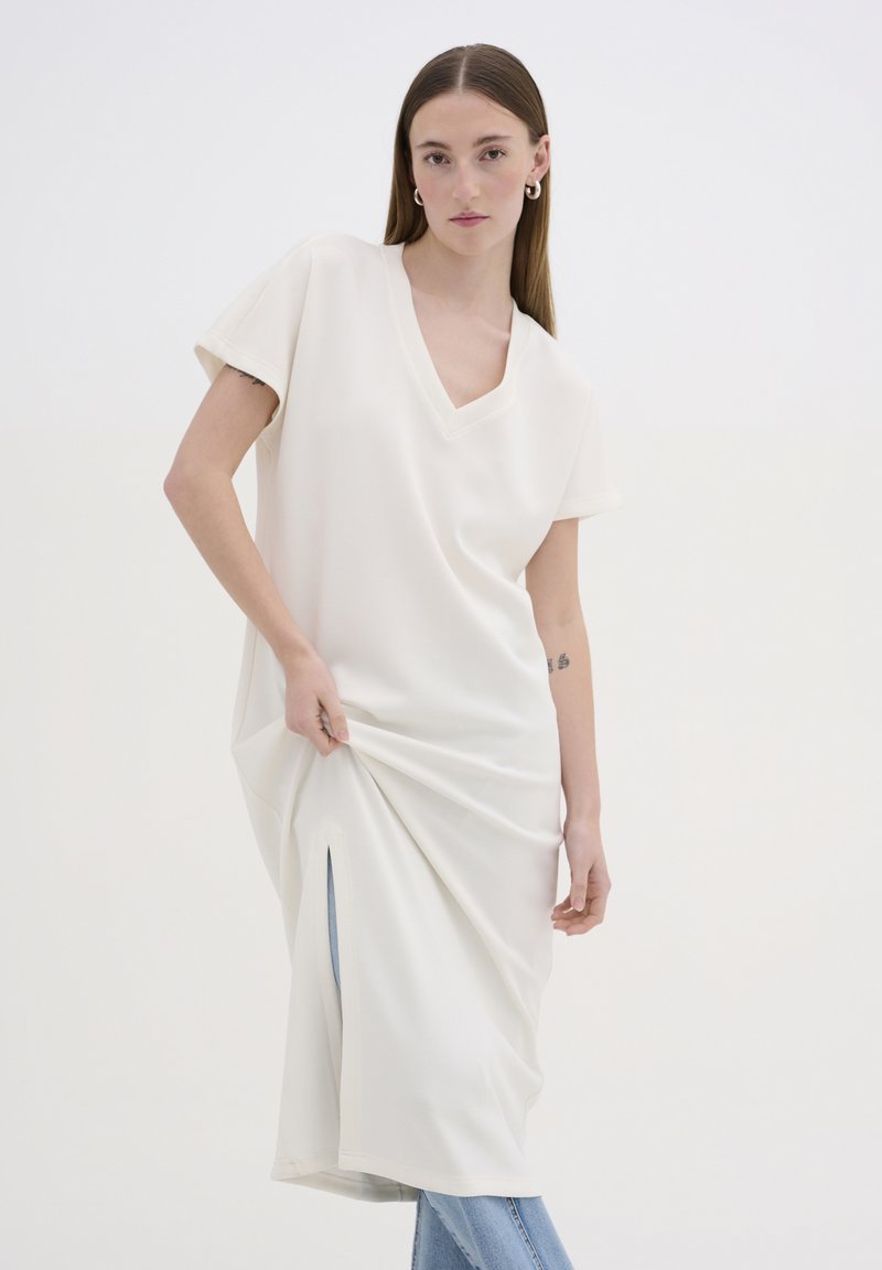 V-hals, kortærmet kjole i blødt, hvidt stof med lige snit og sideslids, parret med lyseblå jeans. Enkel design, minimale detaljer.