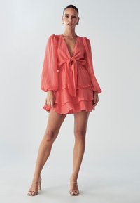 Frau steht in einem korallenroten, langärmligen Kleid mit tiefem V-Ausschnitt und Krawattenfront, kombiniert mit beigen High-Heels.