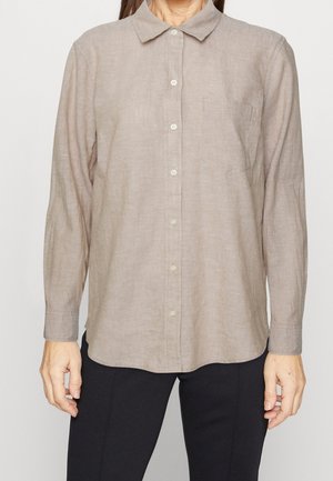 Vrouw draagt een beige blouse met lange mouwen, knopen en kraag, met een borstzak, gecombineerd met een zwarte broek, staand tegen een effen achtergrond.