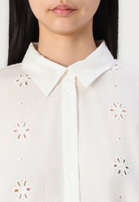Vit linneskjorta med knäppning fram och klassisk krage. Har broderade blommönster längs framsidan för dekorativ detalj.