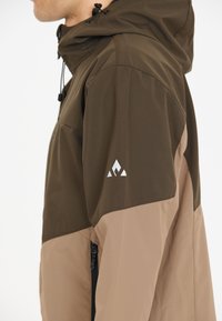 Vandtæt jakke i mørkebrun og beige med en lynlås sidelomme og et hvidt logo på ærmet. Hætte design.