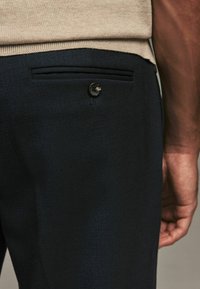 Pantalon bleu marine foncé en tissu texturé, avec une poche arrière dissimulée fermée par un bouton métallique rond, et un gros plan d'une main positionnée à proximité.