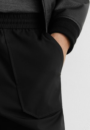 Osoba v černých kalhotách s elastickým pasem a tmavé texturované bundě, s pravou rukou v kapsách kalhot, stojící u bílé zdi.