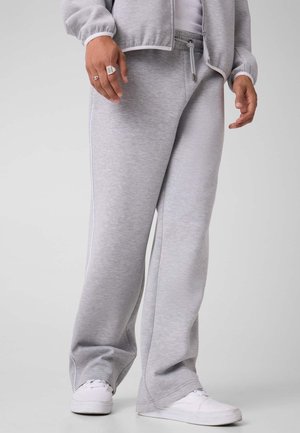 Persona che indossa pantaloni della tuta grigio chiaro, giacca abbinata, scarpe da ginnastica bianche e anelli sulla mano sinistra su uno sfondo uniforme.