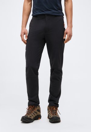 Muž v čiernych outdoorových nohaviciach a hnedých turistických topánkach stojaci na jednoduchom pozadí.