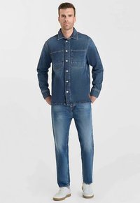 Giacca in denim blu scuro con chiusura a bottoni, due tasche sul petto e colletto appuntito, abbinata a jeans azzurri e sneakers bianche.