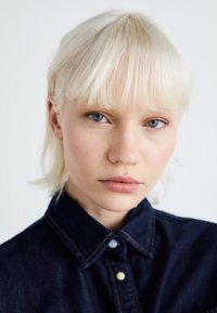 Modrá denimová košeľa s golierom a gombíkovým zapínaním, nosená osobou s priamymi, svetlo blond vlasmi a minimálnym make-upom, na bielom pozadí.