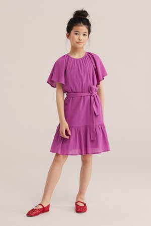 Niña de pie con un vestido morado con mangas con vuelo, un cinturón atado en la cintura y zapatos planos rojos tejidos contra un fondo neutro.