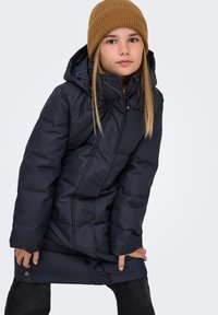 Temno modra podložena jakna s kapuco, z zadrgo spredaj, z gumbi na pritrditev in teksturiranim zaključkom. Oblečena z rjavim rebrastim kapo.