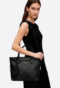 Borsa tote trapuntata nera in ecopelle. Presenta finiture color oro, un dettaglio con nappina e doppi manici con tracolla rimovibile.