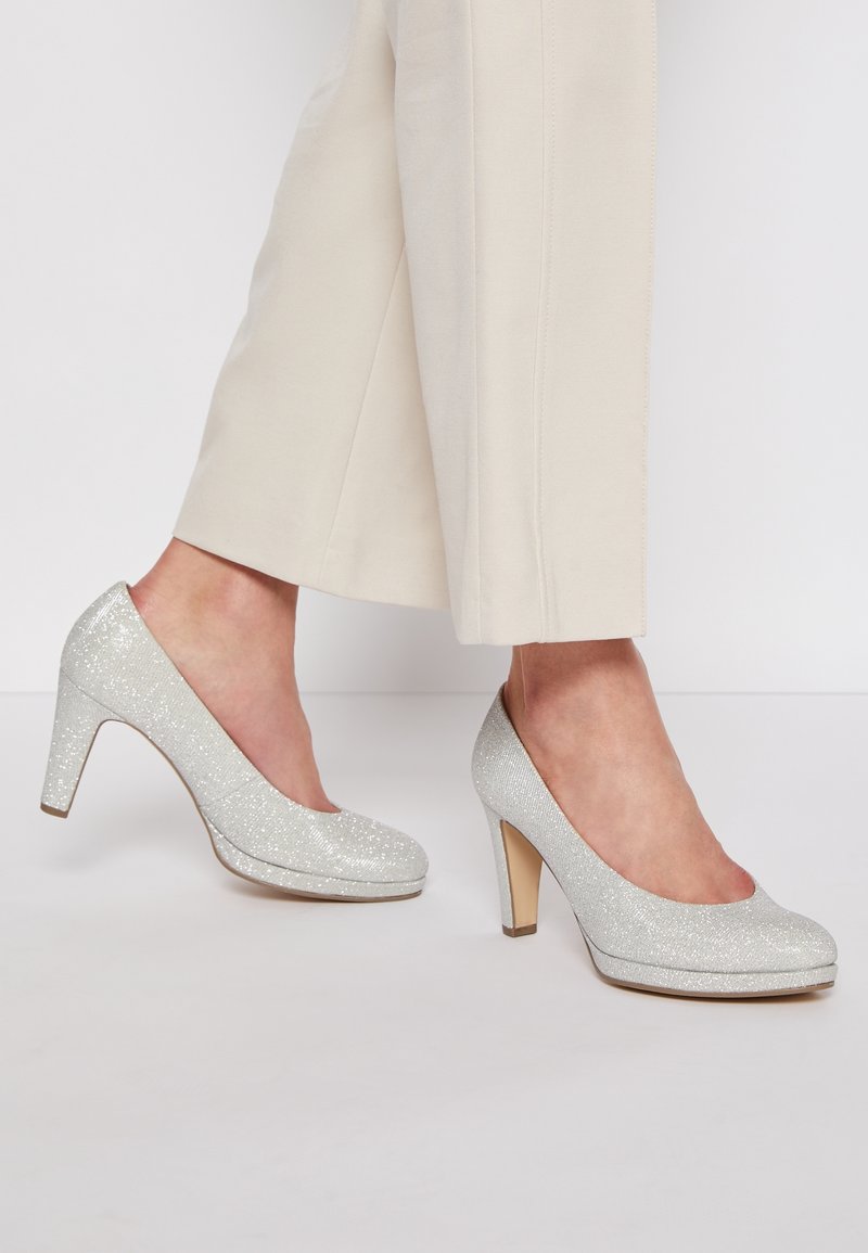 Jambes vêtues d'un pantalon beige et de chaussures à talons hauts argentées scintillantes, posées sur un sol clair devant un fond uni.
