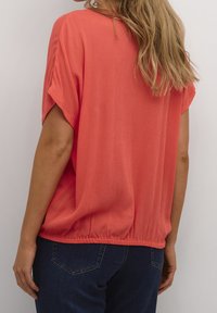 Top corallo ample con maniche corte, tessuto a trama, orlo elastico, abbinato a jeans di denim blu scuro.