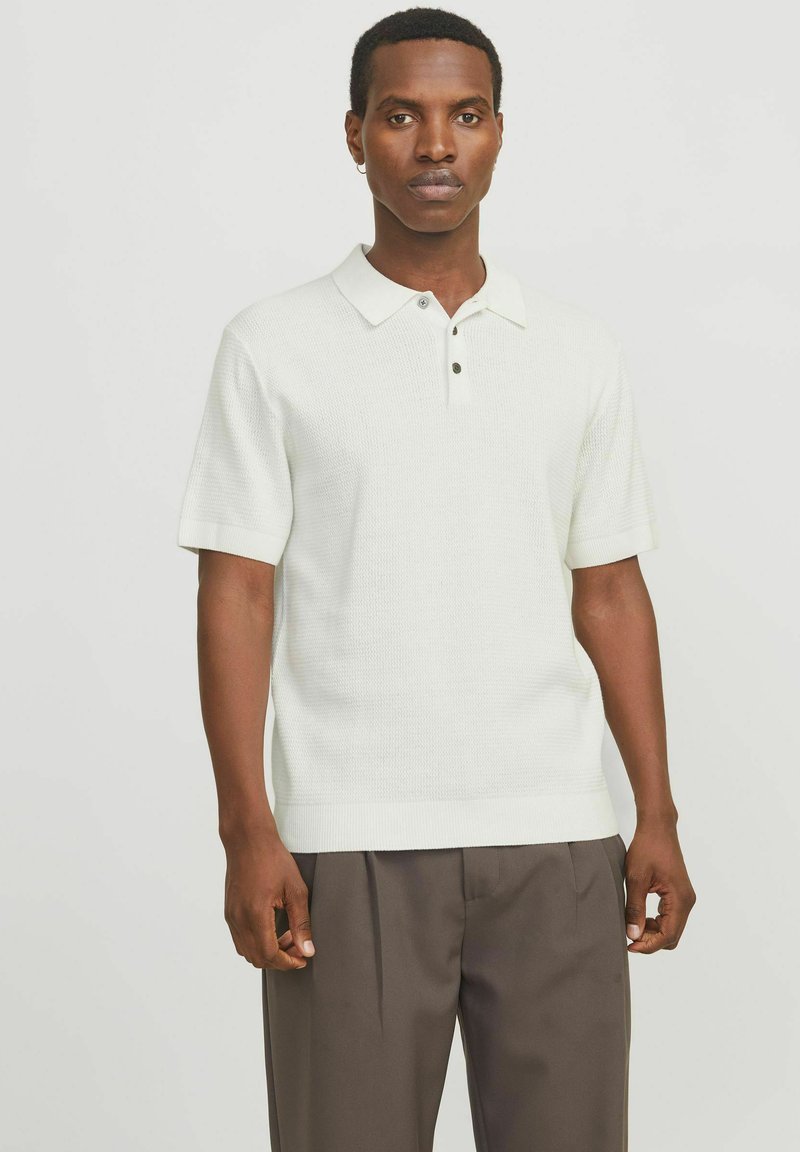 Homme aux cheveux courts portant un polo en maille à manches courtes blanc et un pantalon plissé marron foncé, debout devant un fond uni.