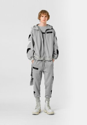 Chándal atlético gris con alas negras bordadas, bolsillos con cremallera y costuras blancas en contraste. Combinado con botas blancas con cordones.