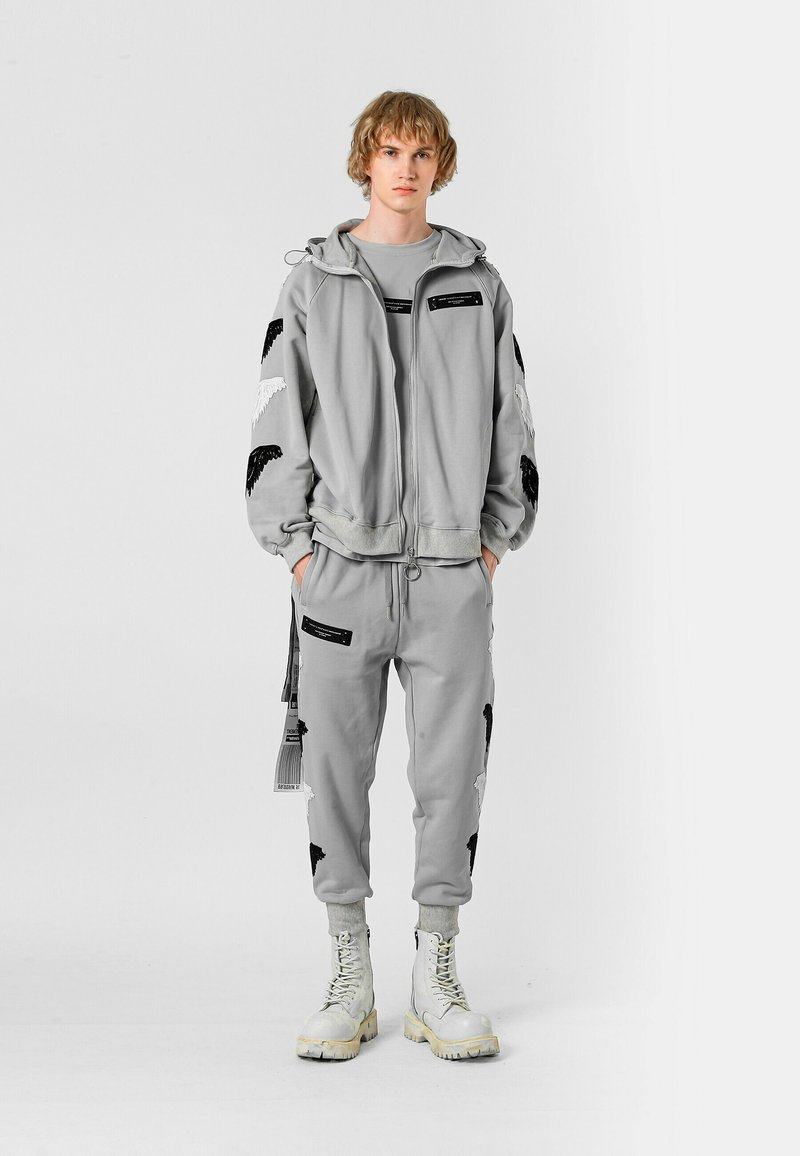Chándal atlético gris con alas negras bordadas, bolsillos con cremallera y costuras blancas en contraste. Combinado con botas blancas con cordones.