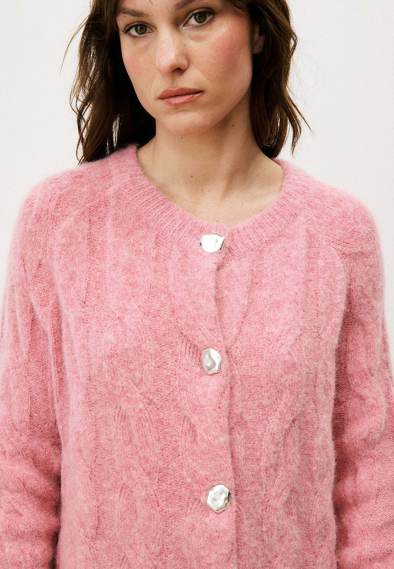Cardigan rose, doux et duveteux, fabriqué en fil moelleux avec un motif texturé ; il présente des boutons ronds argentés brillants et un col côtelé.