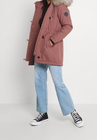 Parka en rosa oscuro con capucha con borde de piel, cintura con cordón y detalles de botones, combinada con vaqueros de pierna ancha en azul claro y zapatillas negras.
