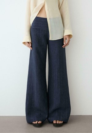 Person, die marineblaue, weite Nadelstreifen-Hosen, ein beiges, transparentes Langarmhemd und schwarze Sandalen trägt, steht vor einem schlichten Hintergrund.