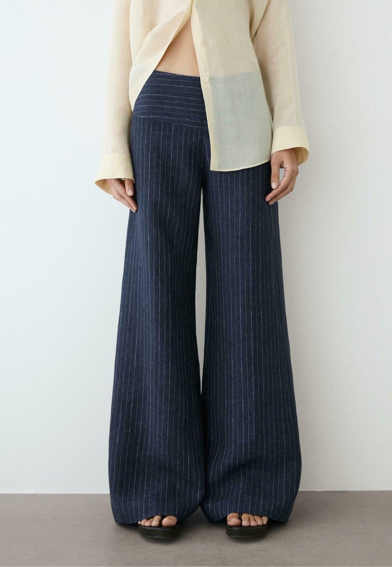 Person, die marineblaue, weite Nadelstreifen-Hosen, ein beiges, transparentes Langarmhemd und schwarze Sandalen trägt, steht vor einem schlichten Hintergrund.