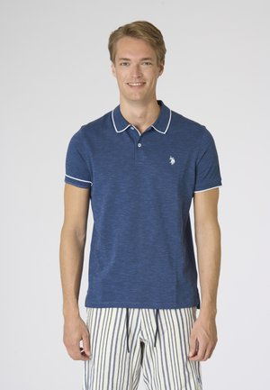 Giovane uomo sorridente, indossa una polo blu a maniche corte con rifiniture bianche e pantaloncini a righe, in piedi davanti a uno sfondo uniforme.