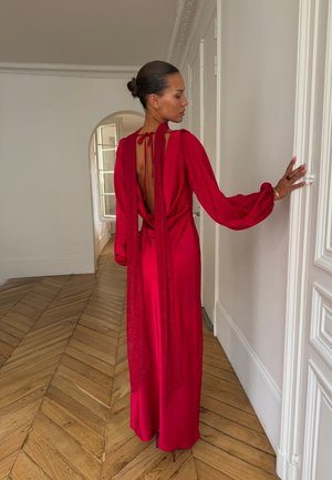 Femme avec les cheveux en chignon portant une longue robe rouge dos nu, debout près d'une porte blanche dans une pièce avec un sol en parquet à chevrons en bois et une fenêtre en arc.