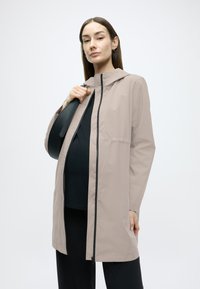 Ανοιχτό μπεζ αδιάβροχο μπουφάν με κουκούλα, με εμπρόσθιο φερμουάρ, πλαϊνές τσέπες και άνετη εφαρμογή, συνδυασμένο με μαύρο τοπ και τσάντα.