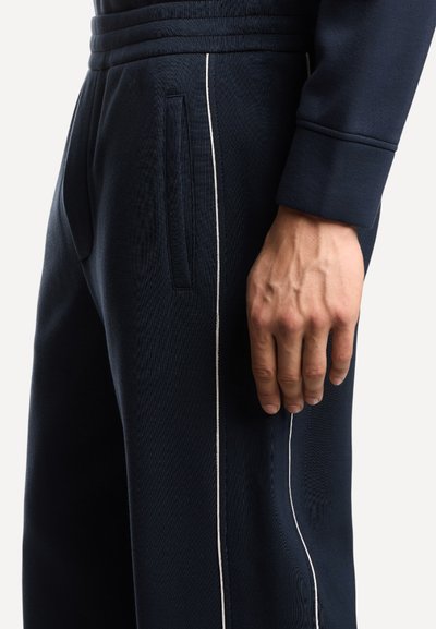Nahaufnahme einer Hand, die neben einer marineblauen Hose mit weißen Seitenstreifen und einer sichtbaren Tasche an der Hüfte ruht.