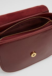 Borsa in pelle bordeaux con pattina in camoscio, hardware dorato e diversi scomparti con zip. Texture liscia con una forma strutturata.