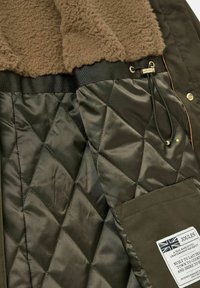 Green quilted jacket interior featuring a brown faux fur lining, elastic waistband, drawstring detail, and a label with care instructions.